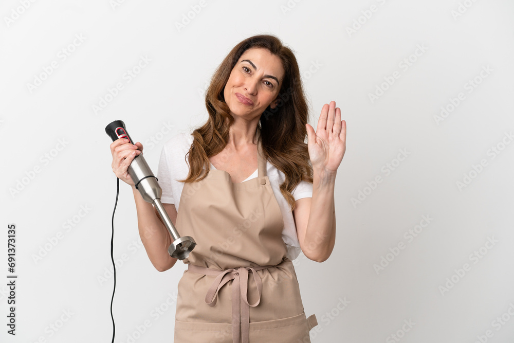 Middle aged caucasian woman using hand blender isolated on white background saluting with hand with happy expression Middle aged caucasian woman using hand blender isolated on white background saluting with hand with happy expression