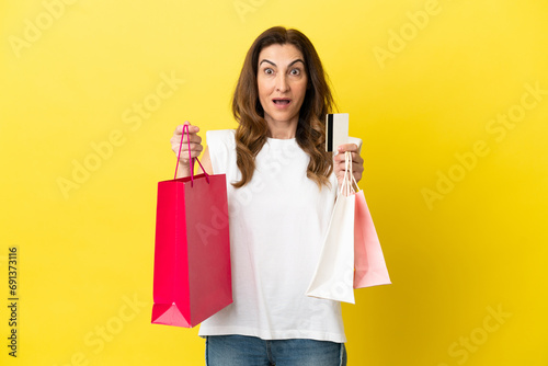 Middle aged caucasian woman isolated on yellow background holding shopping bags and surprised