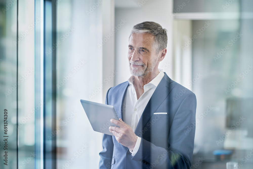 Thoughtful senior businessman standing with tablet PC seen through glass Thoughtful senior businessman standing with tablet PC seen through glass