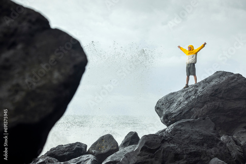 Man with arms raised standing on rock near splashing waves of sea photo