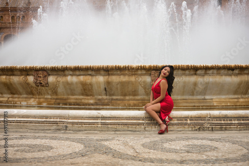 South American woman, young, beautiful, brunette, with an elegant red dress, posing sitting on a fountain in the square of Spain in Seville. Concept of beauty, fashion, ethnicity, diversity, elegance.