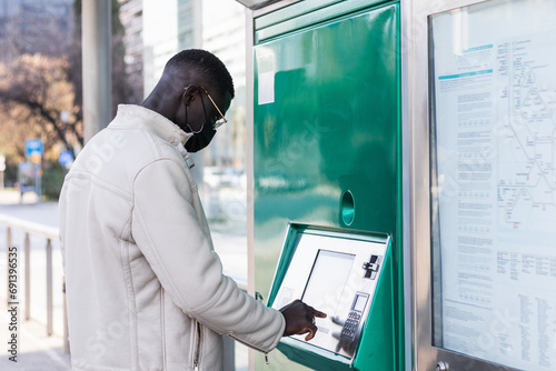 Black man buying a ticket to travel on public transport. He is wearing a medical mask as a protective measure during virus pandemic. photo