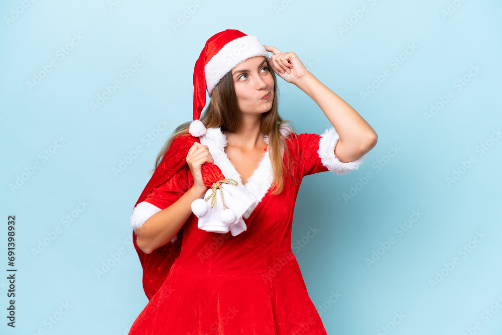 Young caucasian woman dressed as mama noel holding Christmas sack isolated on blue background having doubts and with confuse face expression Young caucasian woman dressed as mama noel holding Christmas sack isolated on blue background having doubts and with confuse face expression