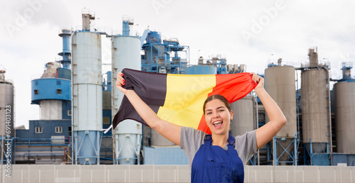 Happy young woman worker holding big flag of Belgium against background of factory photo