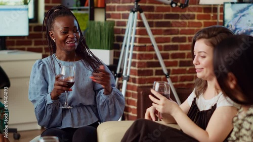 Asian, african american and caucasian women chatting on couch at apartment party, enjoying conversation. Multiethnic BFFs catching up with each other at home, drinking alcohol photo