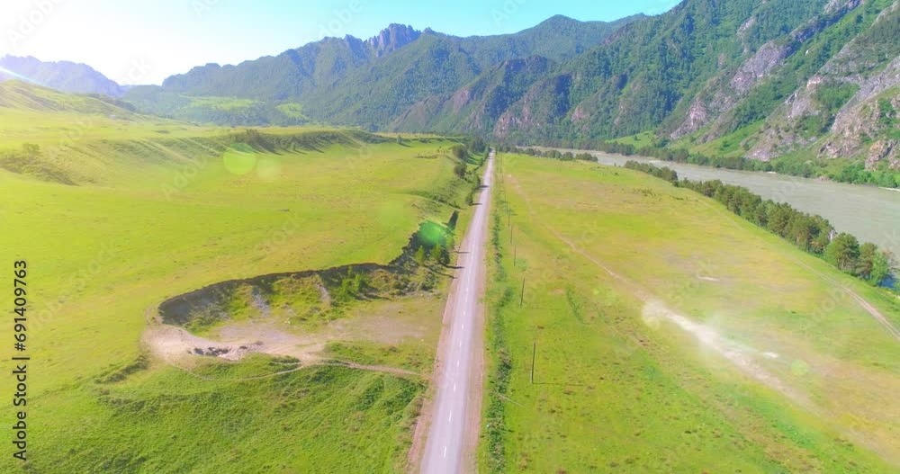 Aerial UHD 4K: mid-air flight over rural mountain road and meadow at sunny summer morning. Green trees and sun rays on horizon. Free rural asphalt highway and river. Aerial UHD 4K: mid-air flight over rural mountain road and meadow at sunny summer morning. Green trees and sun rays on horizon. Free rural asphalt highway and river.