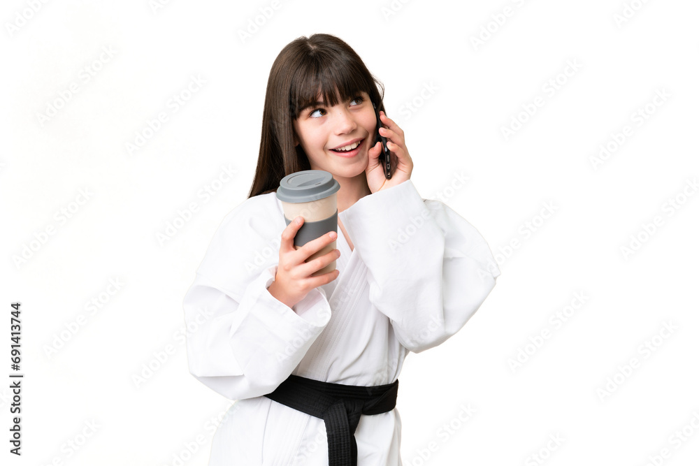 Little Caucasian girl doing karate over isolated background holding coffee to take away and a mobile Little Caucasian girl doing karate over isolated background holding coffee to take away and a mobile