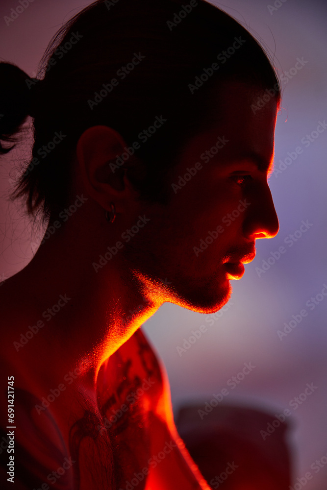 young handsome man with tattoos on body and earring posing in profile surrounded by vibrant lights