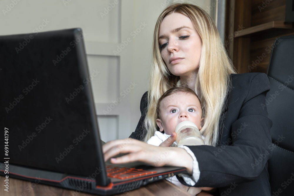 Concept of combining business and caring for a newborn child, young business woman feeds a baby from bottle in office, combining it with work on laptop. Concept of combining business and caring for a newborn child, young business woman feeds a baby from bottle in office, combining it with work on laptop.