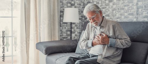 Worried elder senior man feeling bad, upset old middle aged grandfather touching chest feel sudden pain heartburn having heart attack sit on sofa at home. photo