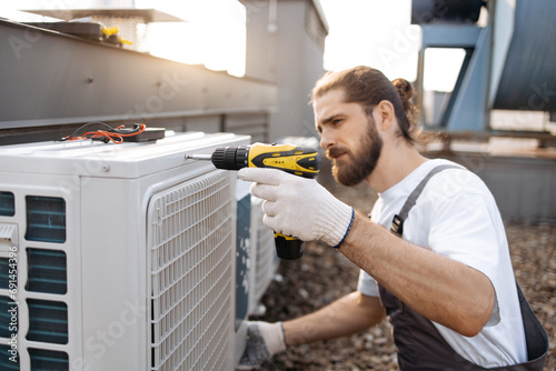 Fotografia Concentrated worker wearing construction gloves screwing bolt into air conditioner using drill in roof of factory