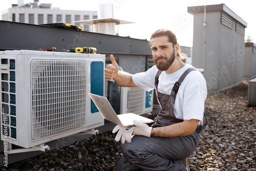 Bearded craftsman in uniform holding digital laptop while checking operation of fan in air conditioner with hand. Caucasian master crouching and using online manual for device outdoors, thumb up.