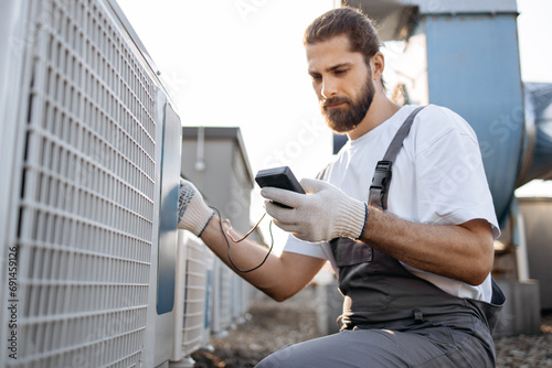 Portrait of bearded worker wearing gray overalls working with air conditioner using voltage tester outdoors. Qualified caucasian man fixing and checking suitability of device on roof of plant. photo