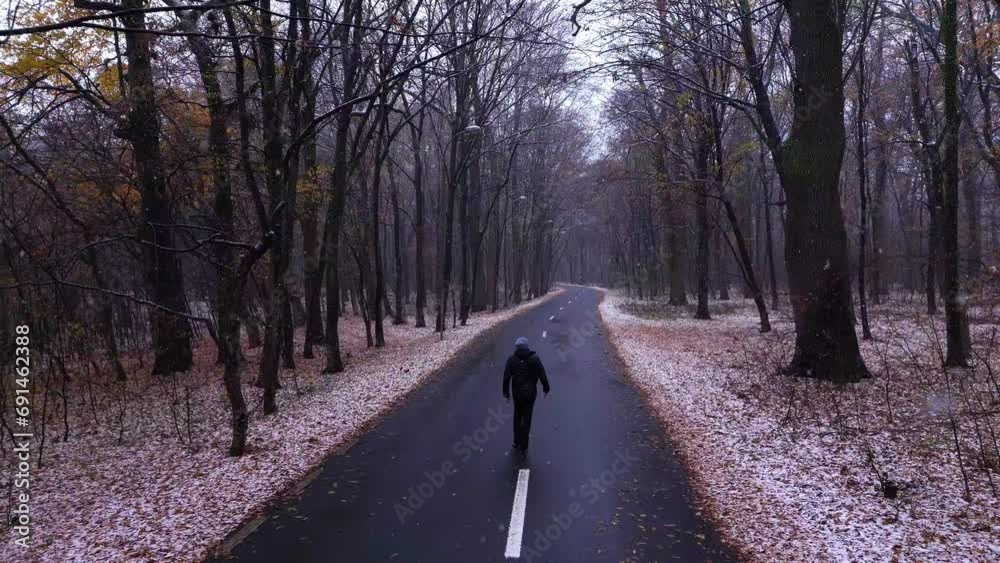 Man in a black jacket walks on the asphalt road in the forest while it is snowing outside. Young Man on the forest road followed by the drone. Aerial view. Autumn weather with snow in the forest.