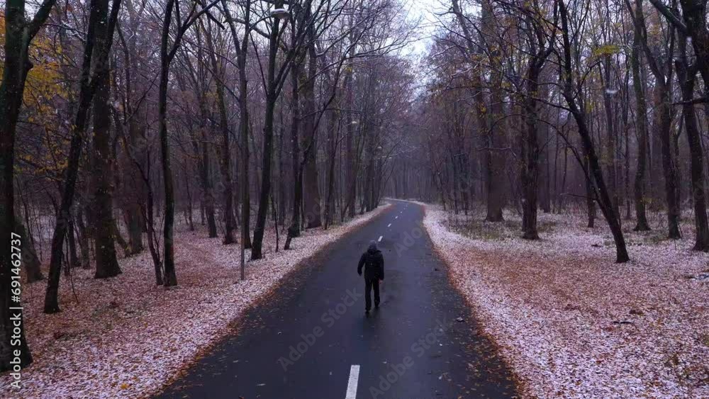Man in a black jacket walks on the asphalt road in the forest while it is snowing outside. Young Man on the forest road followed by the drone. Aerial view. Autumn weather with snow in the forest. Man in a black jacket walks on the asphalt road in the forest while it is snowing outside. Young Man on the forest road followed by the drone. Aerial view. Autumn weather with snow in the forest.