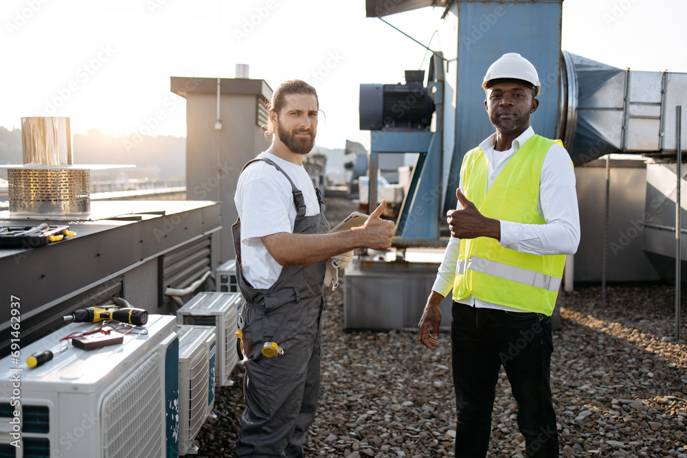 Portrait of positive craftsmen dressed in uniforms standing near air conditioners and smiling while using digital tablet outdoors. Concept of successful cooperation and modern gadgets. Portrait of positive craftsmen dressed in uniforms standing near air conditioners and smiling while using digital tablet outdoors. Concept of successful cooperation and modern gadgets.