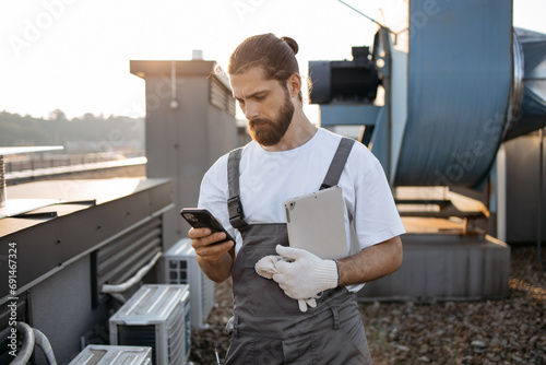 Focused bearded master dressed in overalls standing and holding tablet while using digital smartphone on fresh air. Caucasian skilled man using gadgets and maintaining equipment on roof of factory.
