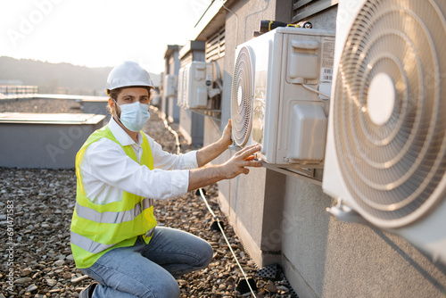 Professional worker of plant wearing dust mask crouching opposite air conditioner and providing inspection on fresh air. Caucasian bearded man checking attachment of device to wall in urban roof.