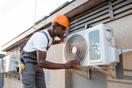 Adult male master wearing gray overalls and orange helmet leaning towards air conditioner and checking fan outdoor. African american man inspecting appliance while repairing in roof of building. photo