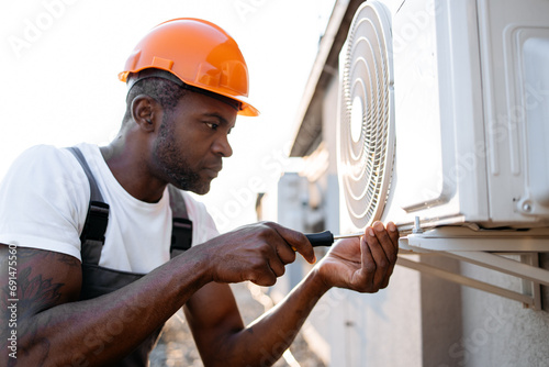 Focused african craftsman wearing orange hard hat holding screwdriver while screwing bolt into air conditioner. Serious specialist repairing broken device and performing work on rooftop.