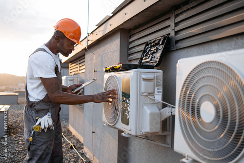 Qualified african american engineer looking at hanging air conditioner on rooftop of residential building. Competent craftsman holding modern tablet and installing device step by step outdoors. photo