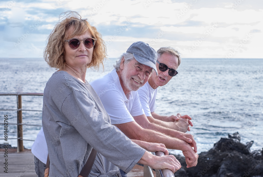 Group of relaxed mature and senior friends standing face the sea at sunset light, three people looking at camera enjoying freedom and vacation or retirement