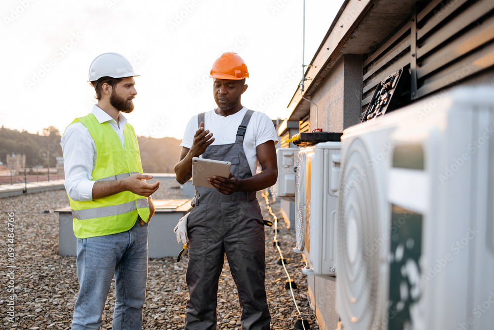 Multiethnic construction managers in protective hard hats reading information from modern tablet about repairing air conditioner. Two men standing on rooftop of industrial factory. Multiethnic construction managers in protective hard hats reading information from modern tablet about repairing air conditioner. Two men standing on rooftop of industrial factory.