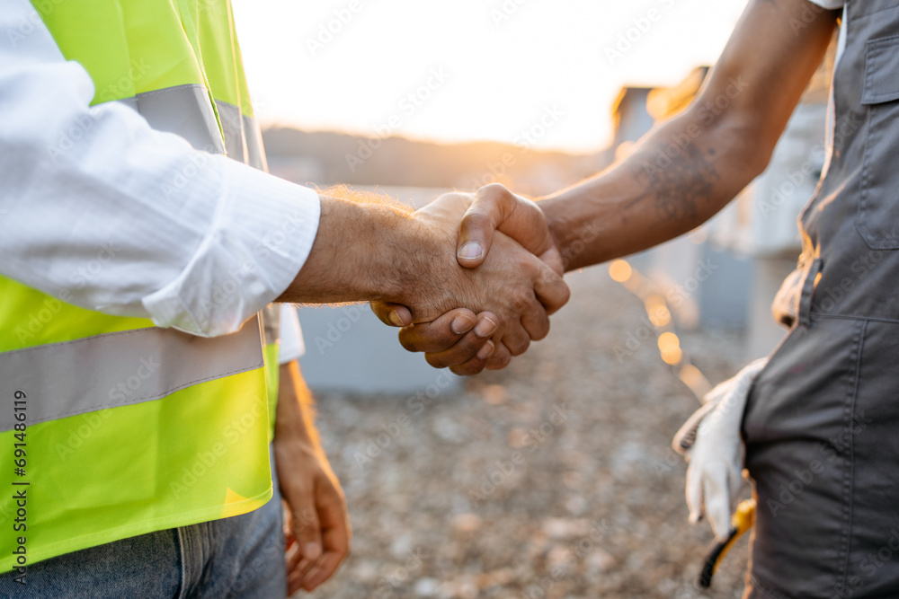 Close up of two strong roof inspectors doing hand shaking while standing during sunset on open air. Responsible male workers concluding deal and agreeing on cooperation together outdoors. - obrazy, fototapety, plakaty 