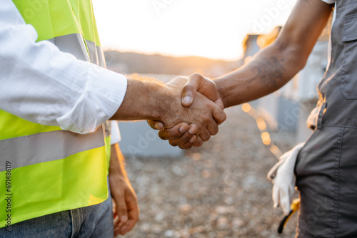 Close up of two strong roof inspectors doing hand shaking while standing during sunset on open air. Responsible male workers concluding deal and agreeing on cooperation together outdoors. photo