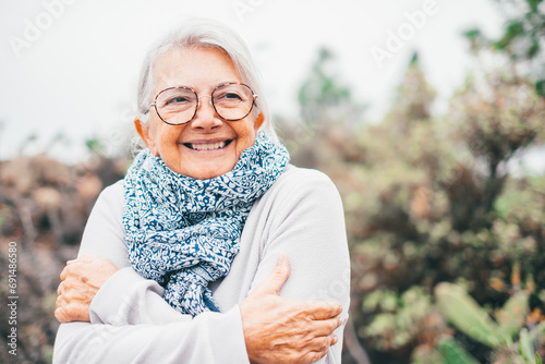 Portrait of attractive smiling senior woman with white hair and eyeglasses embracing herself in a cold foggy day outdoors photo