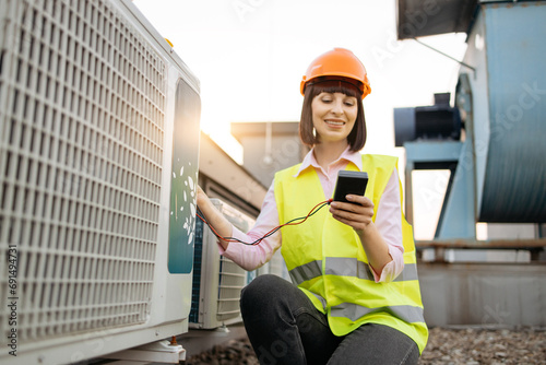 Busy female supervisor wearing orange hard hat and safety vest inspecting air conditioner. Caucasian woman holding voltage tester and taking readings of large climate control unit outdoors. photo