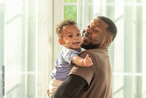 Happy african black parents dad father throw baby son on piggyback and neck riding teasing fun near window. Black baby son and daddy enjoy teasing kiss cheek and neck riding at window light photo