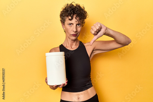 MIddle aged athlete woman holding protein supplement on yellow showing a dislike gesture, thumbs down. Disagreement concept. photo