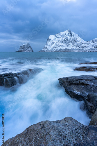 Waves crashing against rocks in the arctic sea with snowcapped mountains in the background