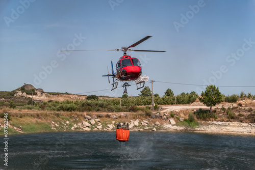 Helicóptero bombeiro a encher o balde que este trás suspenso por baixo num lago para o combate a um incêndio florestal photo