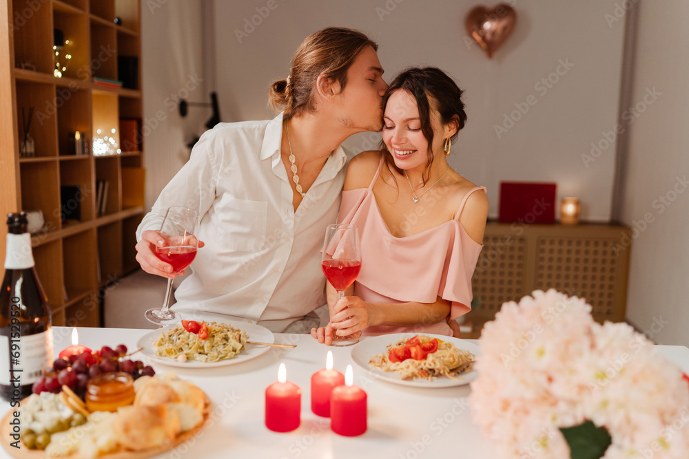 Smiling man kissing a beautiful young woman. Portrait of happy romantic couple drinking wine celebration Valentines day or birthday sitting in restaurant with candles. Dating, love, romantic dinner 