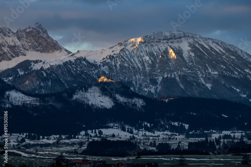 Sunlight falls on Eisenberg castle ruins and Schlo  bergalm in front of an Alpine panorama in the Allg  u