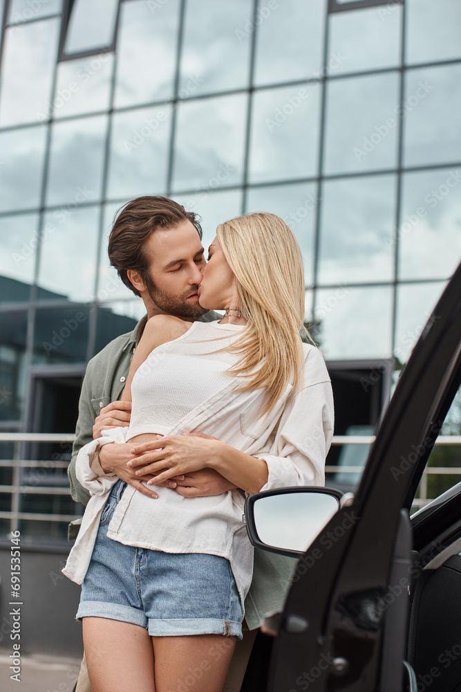 stylish handsome man embracing and kissing blonde girlfriend near modern car on urban street