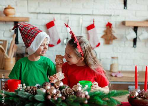 Brother and sister, little boy and girl eating Christmas cookies and drinking drink in Christmas decorations in the kitchen.