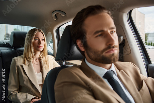 blurred elegant man in formal wear driving luxury car with stylish blonde businesswoman on rear seat