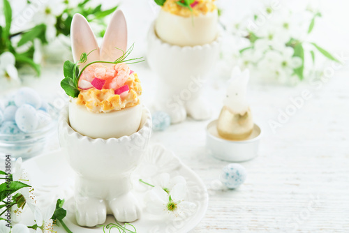 Stuffed or deviled eggs with yolk, shrimp, pea microgreens with paprika in rabbit-shaped stand for easter table decorate fresh cherry or apple blossoms on light background. Traditional dish for Easter photo