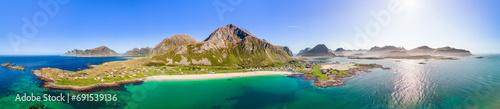 Expansive drone panorama of Jusnesvika Bay and Rambergstranda beach on Flakstadoya island, Lofoten, Norway, with a stunning lens flare over emerald waters, flanked by dramatic peaks