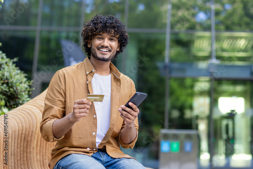 Portrait of a smiling young Muslim man holding a phone and a credit card, sitting outside on a bench and looking at the camera
