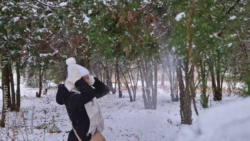 Cheerful young woman in the winter park shaking the snow out of the thuja tree branches, having fun under the snowfall