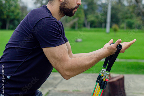 young man training with elastic bands, doing arm exercise outdoors in a park