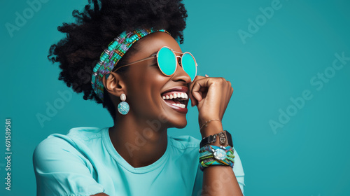 Joyful woman with an afro hairstyle, laughing and wearing turquoise sunglasses against a vibrant turquoise background.