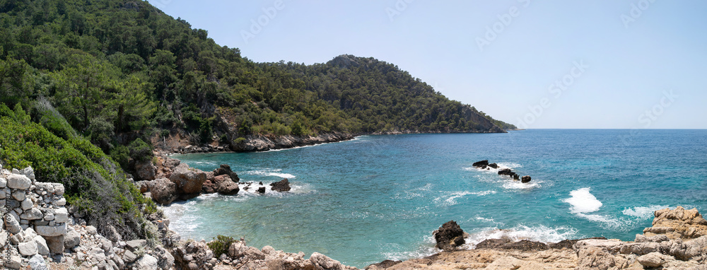 Wide panorama of the rocky beach of Calabantia on the Mediterranean coast of Turkey. Blue lagoon, high rocky cliffs, wooded hills, stone antique ruins