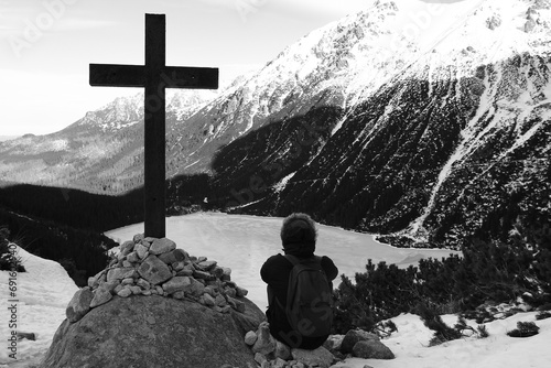 A young man sits near a large cross on a background of mountains and frozen lakes