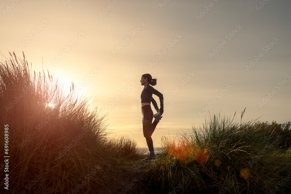 Sporty woman stretching her legs, doing standing quadricep front thigh stretch outdoors at sunrise - obrazy, fototapety, plakaty 