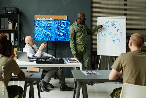 Young African American soldier in camouflage uniform pointing at whiteboard while explaining sketch of military tactics photo